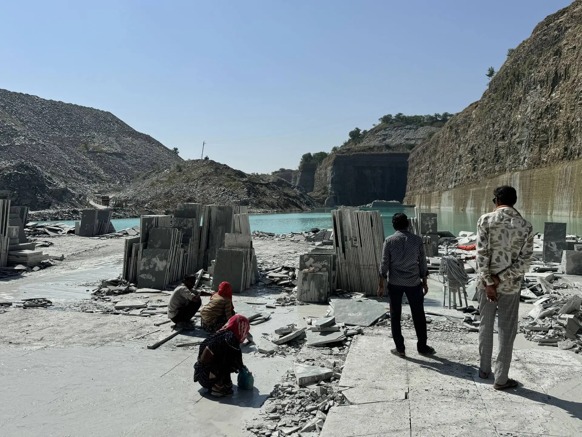Wide view of the quarry pit with stacked slabs, workers and green water below the cut walls
